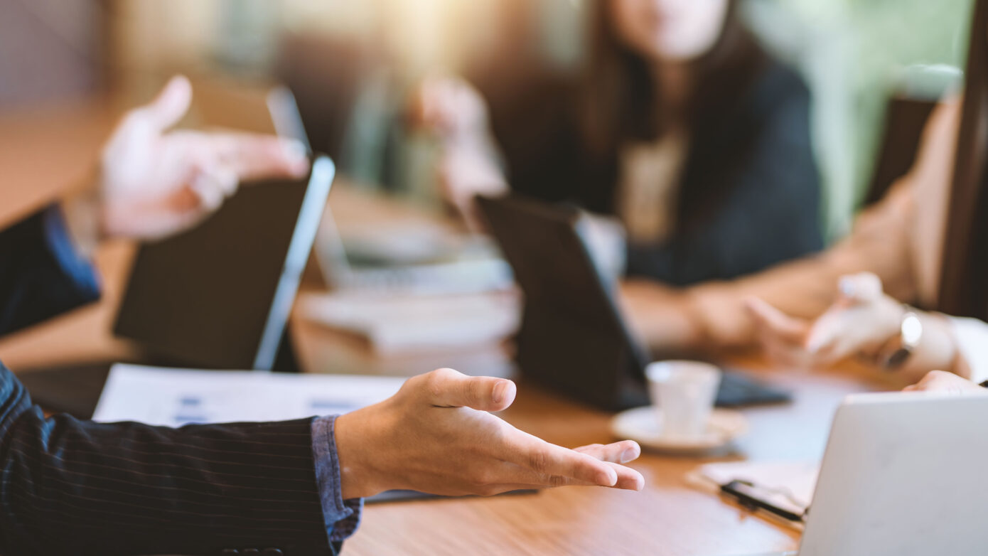 Image of a board room table with smartly dressed nondescript people sitting round it.